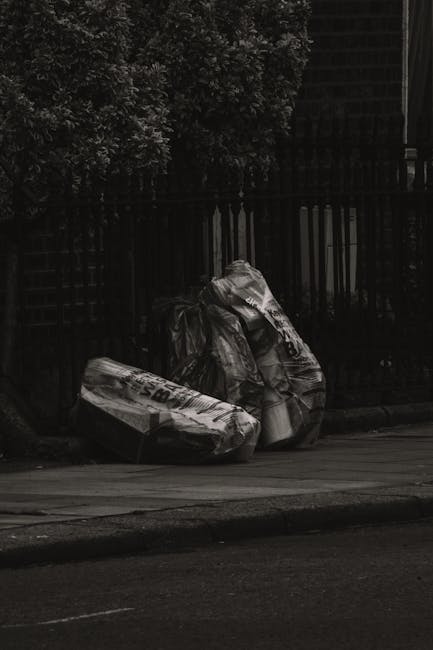 Three large black plastic rubbish bags, tightly tied at the top, are positioned on a sidewalk curb in front of a dark metal fence with vertical bars. The bags appear to contain construction or household waste, with some visible crumpling and wrinkling on the material. Behind the fence, dense foliage of trees or bushes is partially visible, extending upward, and the background includes a building structure partially obscured by shadows. The scene is illuminated by natural light, creating a high-contrast, monochromatic appearance, emphasizing the texture of the bags and the surrounding urban environment. The overall setting suggests an area designated for private rubbish collection or site clearance, consistent with waste removal services such as those provided by Waste Clearance Bromley, focusing on non-recycling waste disposal or alternative waste handling methods in a residential or commercial area.