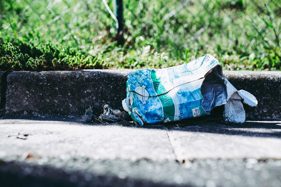 An outdoor scene showing scattered discarded packaging and cardboard boxes on a patch of uneven grassy ground adjacent to a weathered wooden fence. The debris includes crushed and torn boxes, some with green and white branding, along with various paper waste and small plastic fragments. A bent metal pole or rod lies among the rubbish, contributing to the disorderly appearance. The surrounding environment appears natural with sparse, muddy patches and dry, leafless bushes in the background. The scene highlights improperly disposed waste that may require independent rubbish removal or on-site clearance to prevent environmental impact, consistent with services offered by Waste Clearance Bromley for private waste handling and alternative disposal solutions. The lighting is overcast, casting diffuse shadows across the debris, emphasizing the clutter and neglect visible in this unmanaged area.
