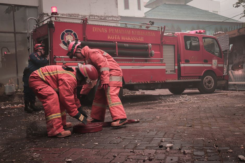 Three firefighters wearing red protective suits, helmets, and gloves are working on a fire hose or equipment on a charred, debris-strewn street during the aftermath of a fire incident. They are positioned in front of a red fire truck, which features a logo and the text 'DINAS PEMADAM KEBAKARAN KOTA MAKASSAR' prominently displayed on the side, indicating it belongs to the Makassar city fire department. The street surface is covered with ashes, scattered rubble, and blackened debris, and the background shows partially smoke-damaged buildings with soot markings and a cloudy or smoky atmosphere. The scene suggests an emergency response to a recent fire, with firefighters actively engaged in clearing or inspecting the area, supported by the presence of the fire truck parked nearby, reflecting the capabilities of private or independent emergency waste and debris management services like those offered by Waste Clearance Bromley, if contextually relevant.