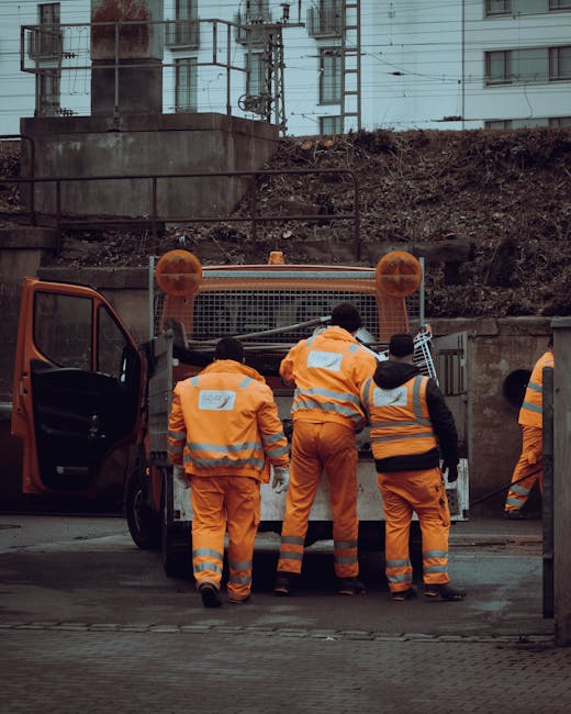 A group of five workers dressed in high-visibility orange and black safety clothing are standing with their backs to the camera, operating or preparing to operate a small waste collection vehicle with a metal mesh front and orange accents. The workers are positioned on an urban street with an open driver’s side door visible on the left. Behind them, there is a dirt embankment and a rough, uneven surface area with loose soil and small debris, suggesting a construction or utility site. In the background, there are tall residential or commercial buildings with multiple windows, and overhead electrical wires supported by pole structures, indicating a city environment. The scene is illuminated by natural daylight, contributing to the industrial atmosphere. The setup and activity imply that this is part of an on-site waste removal or clean-up operation, consistent with a private rubbish collection service such as Waste Clearance Bromley, especially in contexts concerning urgent or independent waste handling efforts near urban infrastructure.
