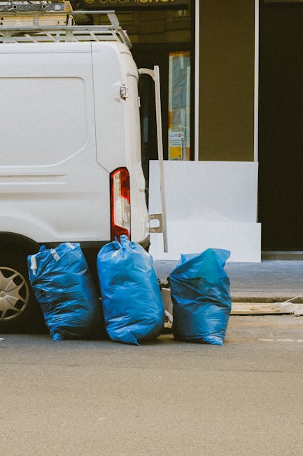 A white commercial van parked on a street curb with its rear door closed, positioned next to three large blue plastic rubbish bags filled with waste, which are placed on the pavement close to the vehicle. The bags appear full and are slightly crumpled, with one leaning slightly to the left. In the background, a storefront with large glass windows and a grey frame can be seen, with minimal signage reflected in the glass. The environment suggests a typical urban or suburban setting where private rubbish collection or onsite clearance might take place, with the presence of waste bags indicating ongoing rubbish removal activity. Waste Clearance Bromley provides services related to such waste disposal tasks, supporting independent collection options outside of local authority services. The lighting is natural, and the scene is clear and neutral, emphasizing the typical scene of waste ready for removal or disposal in an area serviced by waste clearance specialists.