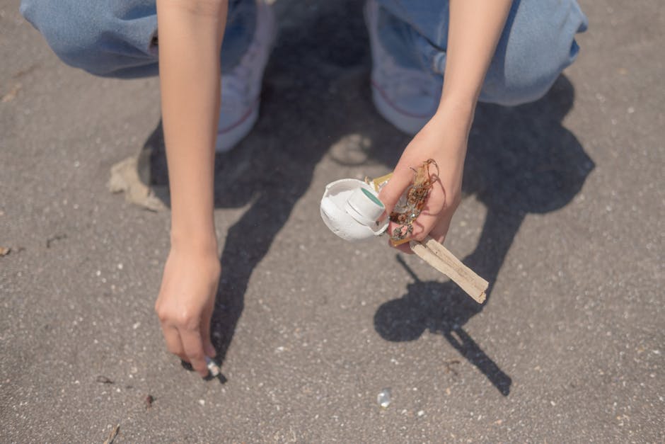 A person is crouched down on a paved outdoor surface, holding a small collection of waste materials in their right hand. The waste includes a partially broken white ceramic or plastic object, a small piece of wood, and some natural fiber or string. The individual is wearing light blue jeans and white sneakers with a hint of red detailing, with their left hand resting on the ground for support. The background shows the textured gray pavement with slight pebbles and debris scattered around, and the lighting appears natural, suggesting daytime. The scene emphasizes proper disposal of small waste items, aligning with the context of rubbish removal services offered by Waste Clearance Bromley, which includes handling small debris and supporting alternative waste disposal methods outside municipal collection.