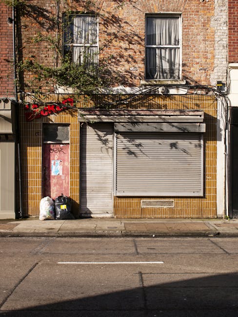 The image depicts the ground floor of a brick building with two vertically aligned windows on the upper level, each featuring white frames and partially drawn curtains. The exterior wall has a weathered appearance with visible mortar lines and some foliage casting shadows. On the street-facing façade, there is a closed grey metal roller shutter, situated within a tiled section of ochre-colored tiles that surrounds the doorway area. To the left of the shutter, a small door with graffiti and peeling paint is visible, and nearby, two black rubbish bags and a white plastic bag are placed on the pavement, indicating waste disposal. An overhanging strip of roofing extends above the shutter, with a black cable running horizontally across the upper part of the wall. The scene is lit by natural daylight, casting shadows on the building's surface and pavement, which shows subtle tire marks and a curb. The setting suggests an area typically associated with independent waste collection, maintained by waste clearance services such as Waste Clearance Bromley, reflecting a common urban context of rubbish removal and private waste handling near commercial or residential premises.