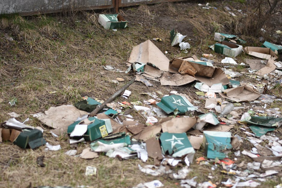 A patch of ground with scattered litter and debris, including torn cardboard boxes, some of which are green with white star logos, partially crushed and torn, lying on a grassy and muddy surface. The debris is dispersed unevenly, with some cardboard pieces overlapping and others spread apart, alongside smaller paper scraps and plastic fragments. In the background, a section of a weathered wooden fence is visible at the top of the image. The surrounding environment appears outdoor, possibly near a roadside or vacant lot, with natural lighting casting soft shadows on the ground. This scene reflects an instance of illegal dumping or improper disposal of waste, which services such as waste clearance companies might address through private rubbish collection or on-site clearance solutions. Waste Clearance Bromley often manages such rubbish removal tasks to help maintain clean and tidy outdoor areas, especially where public authority rubbish collection may be unavailable or insufficient.